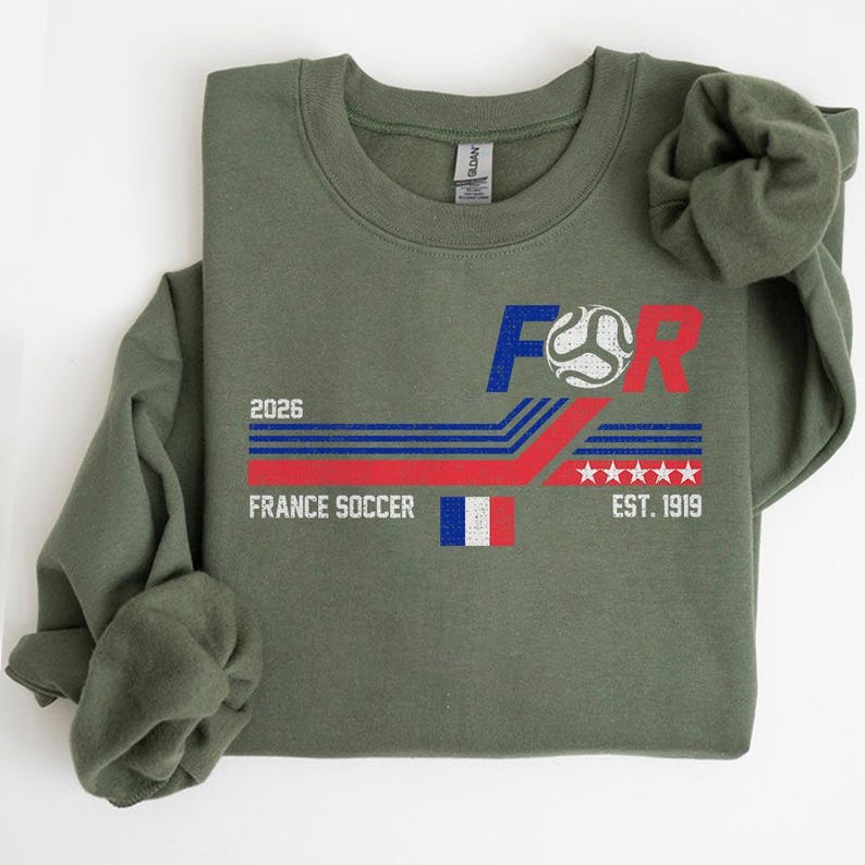 May include: Olive green sweatshirt with a graphic design for France Soccer. The design includes the text "FOR", a soccer ball, the French flag, the year "2026", and "EST. 1919". The design features red, white, and blue colors.