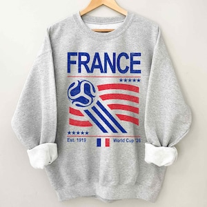 Pu&ograve; includere: Una felpa grigia con la scritta "FRANCE" in blu. Sotto, un disegno di pallone da calcio con strisce rosse e blu, e il testo "Est. 1919" e "World Cup '26". La felpa ha polsini bianchi.