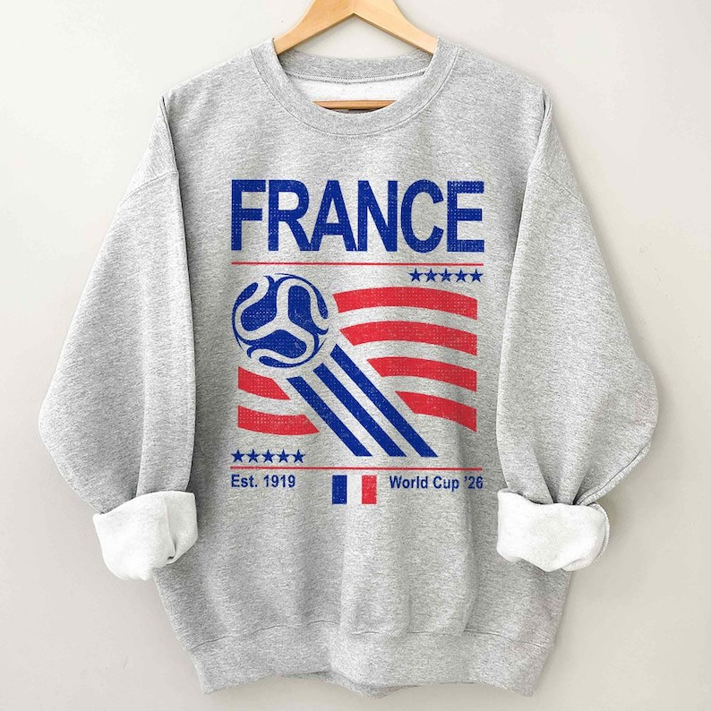 Pu&ograve; includere: Una felpa grigia con la scritta "FRANCE" in blu. Sotto, un disegno di pallone da calcio con strisce rosse e blu, e il testo "Est. 1919" e "World Cup '26". La felpa ha polsini bianchi.
