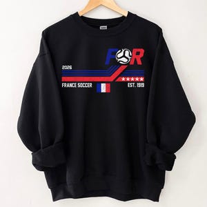 May include: Black sweatshirt with a graphic design featuring the text "FOR" above a soccer ball, with "2026" and "FRANCE SOCCER" below. The design includes the French flag and "EST. 1919". The sweatshirt is hanging on a wooden hanger.