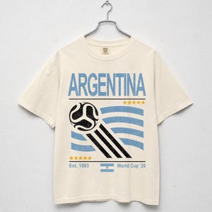 May include: Cream-colored t-shirt with the word "ARGENTINA" in light blue block letters. Below the text is a soccer ball graphic with blue and black stripes, and the text "Est. 1893" and "World Cup '26".