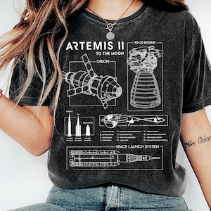 Puede incluir: Camiseta gris oscuro con un gráfico blanco detallado de la misión Artemis II a la luna, incluyendo la nave espacial Orión, el motor RS-25 y diagramas del sistema de lanzamiento espacial. La camiseta tiene un aspecto vintage.