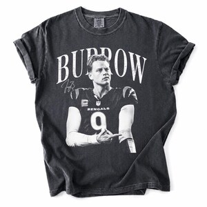 May include: A charcoal grey short-sleeve t-shirt featuring a black and white graphic of Joe Burrow in a Bengals jersey with the number 9. The text BURROW is printed in large white letters above the image, accompanied by a signature graphic.