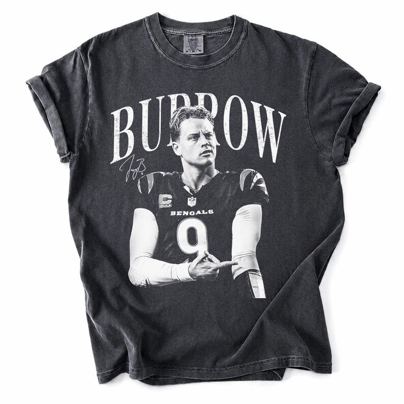 May include: A charcoal grey short-sleeve t-shirt featuring a black and white graphic of Joe Burrow in a Bengals jersey with the number 9. The text BURROW is printed in large white letters above the image, accompanied by a signature graphic.