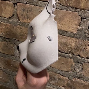 May include: A white animal mask with black painted details. The mask is made of a smooth, pliable material and has a three-dimensional shape. The mask has a large, round eye opening on the left side and a smaller, oval eye opening on the right side. The mask also has a small, black nose and a black mouth.