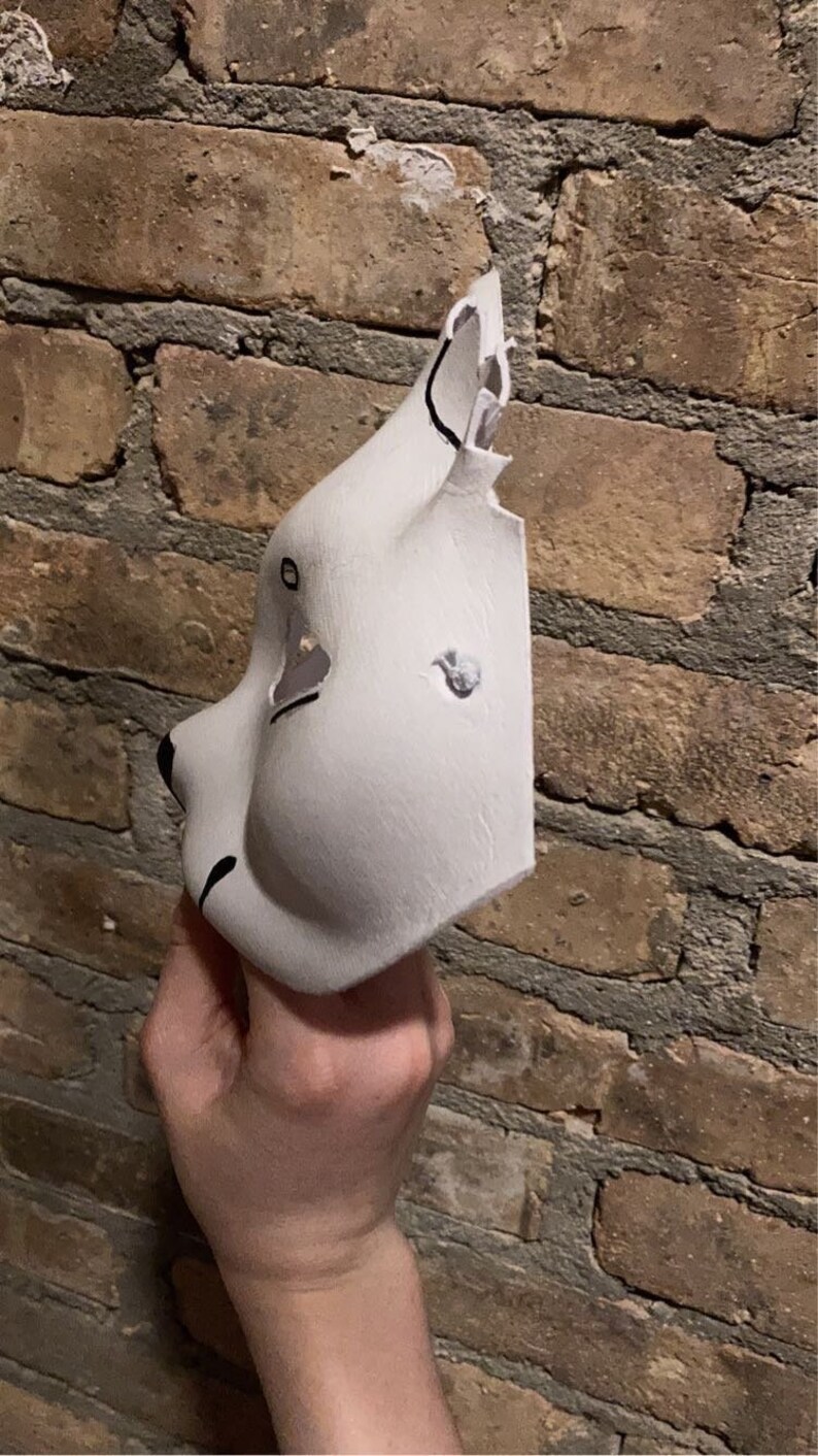 May include: A white animal mask with black painted details. The mask is made of a smooth, pliable material and has a three-dimensional shape. The mask has a large, round eye opening on the left side and a smaller, oval eye opening on the right side. The mask also has a small, black nose and a black mouth.