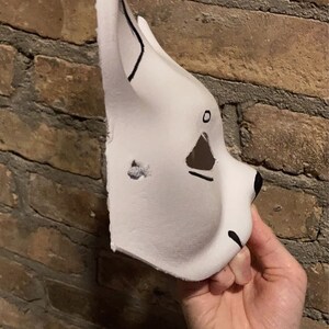 May include: A white furry mask with black painted eyes and nose. The mask is made of foam and has a simple design.