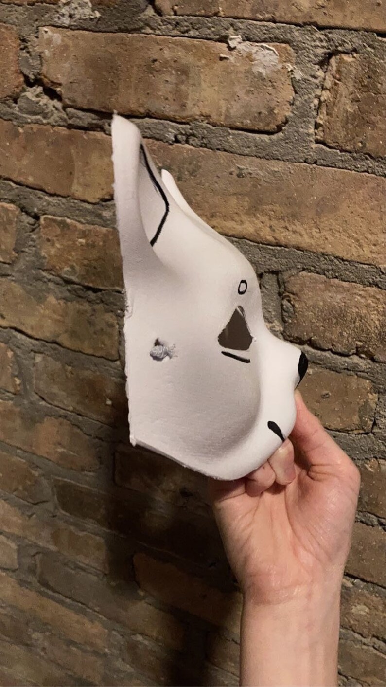 May include: A white furry mask with black painted eyes and nose. The mask is made of foam and has a simple design.