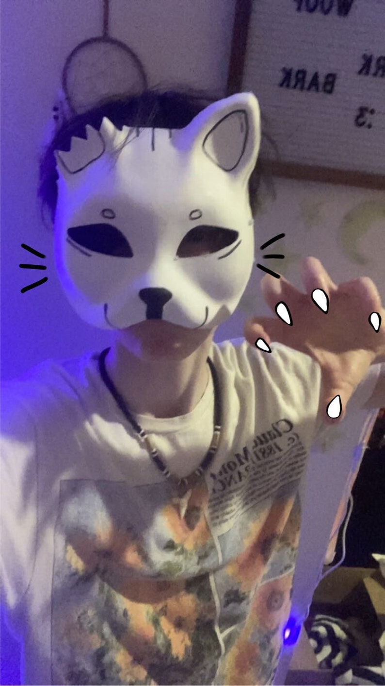 May include: A person wearing a white cat mask with black markings. The mask has a black nose and two black eyes. The person is wearing a white shirt with a floral print and a black beaded necklace.