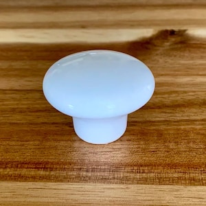 May include: A white, mushroom-shaped cabinet knob with a rounded top and a short, cylindrical base. The knob is made of a smooth, glossy material and is set against a warm, wooden background. This hardware is suitable for drawers and cabinets.