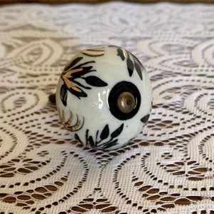 May include: White ceramic knob with black and gold floral design. The knob has a black center with a gold accent.