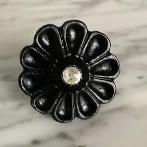 May include: A black, flower-shaped cabinet knob with a clear, round crystal center. The knob has a glossy finish and is set against a marble surface. This decorative hardware is suitable for drawers and cabinets.