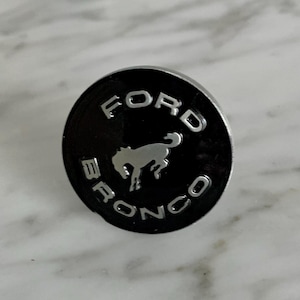 May include: A black and silver Ford Bronco emblem with a stylized horse logo.