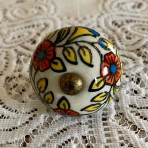 May include: A round, white ceramic cabinet knob with a brass screw. The knob is decorated with colorful floral designs in orange, yellow, and blue, with green leaves. The knob is a decorative hardware element.