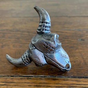 May include: A silver-colored bull skull figurine with curved horns and detailed texture. The sculpture is set against a wooden surface, showcasing the intricate design of the bovine skull.