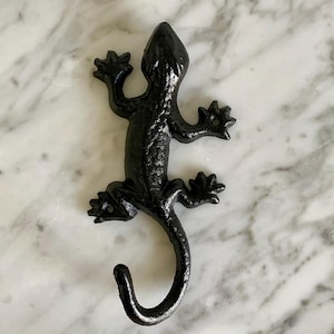 May include: A black metal lizard-shaped wall hook. The hook is designed to resemble a gecko, with detailed legs, a textured body, and a curved tail. The hook is approximately 6 inches long.