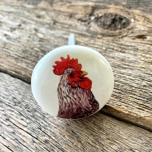 May include: White ceramic knob with a rooster illustration. The rooster has a red comb and wattle, gray and white feathers, and a brown body.