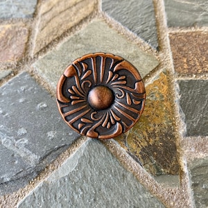May include: A copper-toned cabinet knob with a decorative floral design. The knob has a raised center and a circular shape.