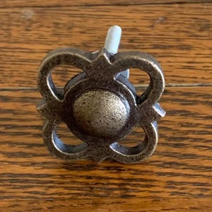 May include: A bronze-colored cabinet knob with a decorative floral design. The knob features a central circular element surrounded by four symmetrical, openwork petal shapes. A threaded post extends from the back for attachment.