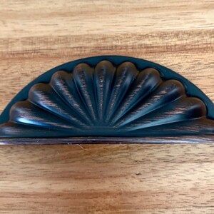 May include: A decorative, half-moon cabinet pull with a dark bronze finish. The pull features a fan-like design with a dark teal backing. The hardware is designed to be mounted on a cabinet or drawer.