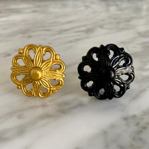 May include: Two decorative cabinet knobs. One is gold-colored, the other is black. Both knobs have a floral design with a central circular element and curved, openwork petals. The knobs are displayed on a marble surface.