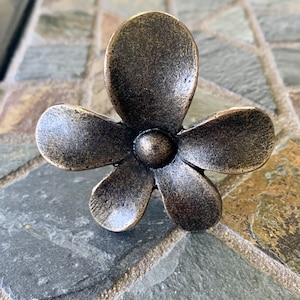 May include: A bronze-colored flower-shaped cabinet knob. The knob has five petals and a small, round center. The finish has a slightly aged appearance. The knob is designed for use on drawers or cabinets.
