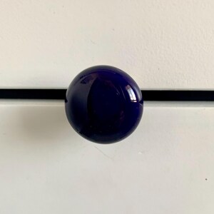 May include: A round, dark blue glass cabinet knob. The knob is a sphere shape and has a glossy finish. The knob is mounted on a white surface with a black horizontal accent line.