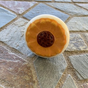 May include: A round, decorative cabinet knob with a cream-colored base and a painted design. The design features a central dark brown circle surrounded by concentric rings of orange and yellow, resembling a stylized flower or fruit. The knob is likely ceramic.