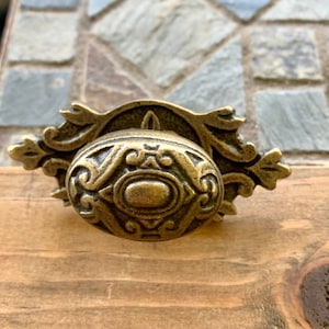 May include: A vintage-style, ornate brass cabinet knob with a detailed oval design. The knob has a decorative backplate with a floral pattern.