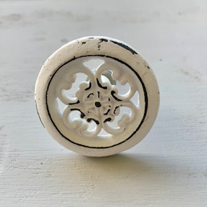 May include: White cast iron cabinet knob with a decorative, ornate design. The knob has a distressed finish and a black outline.