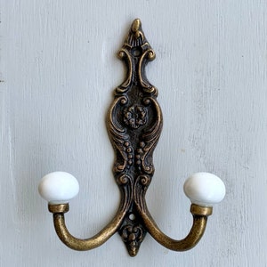 May include: Ornate double hook with a decorative scroll design in an antique brass finish. The hooks have white ceramic finials.