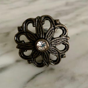 May include: Bronze-toned, floral-shaped cabinet knob with a clear, faceted crystal center. The knob features an ornate, openwork design with stylized leaves and curved elements, suitable for furniture or decor.