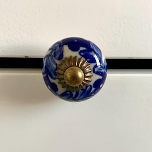 May include: A round cabinet knob with a blue and white floral design. The knob has a brass-colored base and is mounted on a white surface. The knob is a decorative element for furniture.