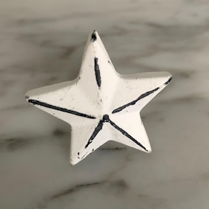 May include: White star-shaped decorative object with black accent lines. The star has a distressed, vintage appearance. The object is likely made of metal or a similar material. The star is a decorative element.