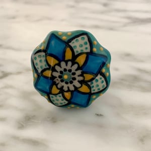 May include: A decorative ceramic cabinet knob with a floral design. The knob features a turquoise base with a blue, yellow, and white flower pattern. Small yellow dots are scattered around the edges. The knob is likely used for furniture.
