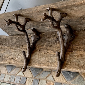 May include: Two rustic brown cast iron hooks shaped like branches with multiple prongs. The hooks are mounted on a weathered wooden surface.