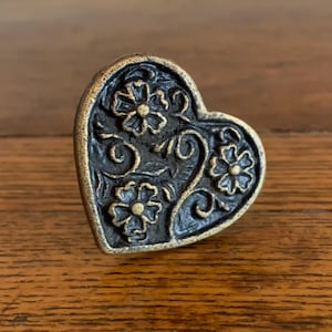 May include: A heart-shaped cabinet knob with an antique brass finish. The knob features an embossed floral design with three flowers and swirling vines. The background is a dark, contrasting color, enhancing the details of the design. The knob is likely made of metal.