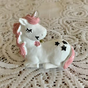 May include: A white unicorn figurine with pink mane, tail, and accents. The unicorn has a silver horn, black star details, and closed eyes. The figurine is resting on a white lace surface.