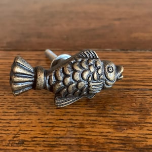 May include: A bronze-colored fish-shaped cabinet knob. The knob features detailed scales, fins, and a tail. The hardware is mounted on a silver post. The knob is designed to add a decorative touch to furniture.