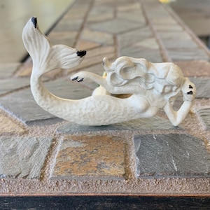 May include: White ceramic mermaid figurine with black accents. The mermaid is lying on her side with her tail curled up behind her.