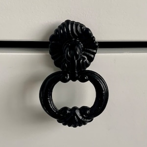 May include: Black metal drawer pull with a decorative, ornate design. The pull has a ring shape with a detailed, floral-like top.
