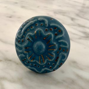 May include: A round, teal ceramic cabinet knob with a floral design. The knob features a central starburst pattern in gold, surrounded by intricate detailing and small brown dots. The knob is set against a marble surface.