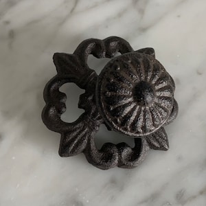 May include: A black cast iron cabinet knob with a decorative floral design. The knob has a round, raised center with a sunburst pattern.