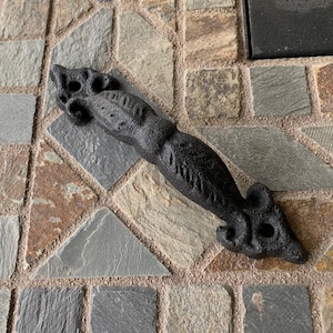 May include: A black cast iron door handle with a decorative design. The handle is shaped like a stylized animal, possibly a mouse or rat, with a textured surface and visible bolt holes. The handle is lying on a multi-colored stone surface.
