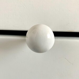 May include: A white, spherical cabinet knob. The knob is glossy and reflects light, suggesting a ceramic or porcelain material. It is mounted on a white surface with a black accent line.