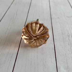 May include: A faceted, peach-colored glass cabinet knob with a silver metal screw. The knob has a flower-like design with multiple facets. The background is a light-colored wooden surface.
