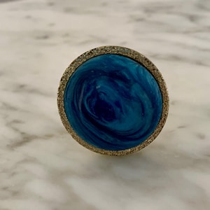May include: A round, decorative cabinet knob with a swirling blue and teal center. The knob is framed by a textured, gold-colored metal. The design is reminiscent of ocean waves.