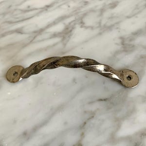 May include: A silver-toned, twisted metal cabinet pull with a curved design. The handle has a textured, rope-like appearance and features circular mounting plates with screw holes on each end. This hardware is suitable for furniture restoration or DIY projects.