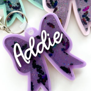 Puede incluir: Llavero de lazo de resina p&uacute;rpura con el nombre "Addie" en letras blancas. El lazo tiene un acabado brillante e iridiscente.