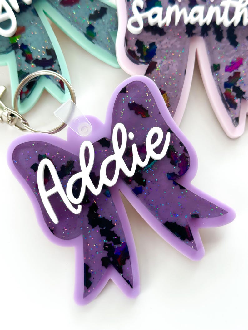 Puede incluir: Llavero de lazo de resina p&uacute;rpura con el nombre "Addie" en letras blancas. El lazo tiene un acabado brillante e iridiscente.